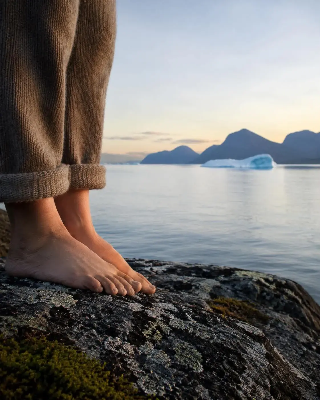 Bare fødder på en klippe ved vandet med grønlandske fjelde og et isfjeld i baggrunden